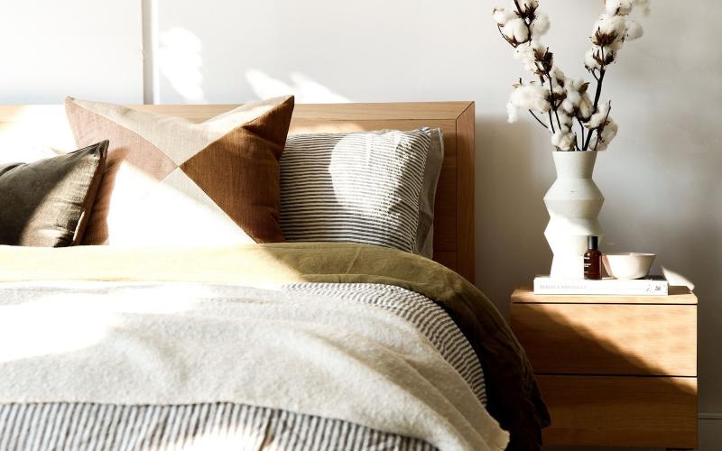 swath of sunlight shines across a well made bed and bedside table with a vase of cotton plants